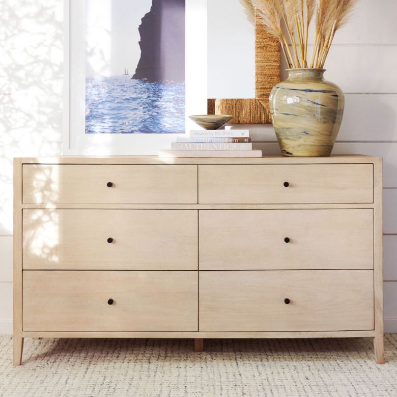 Light wood dresser with six drawers, decorated with books, a ceramic vase of pampas grass, and framed art above.