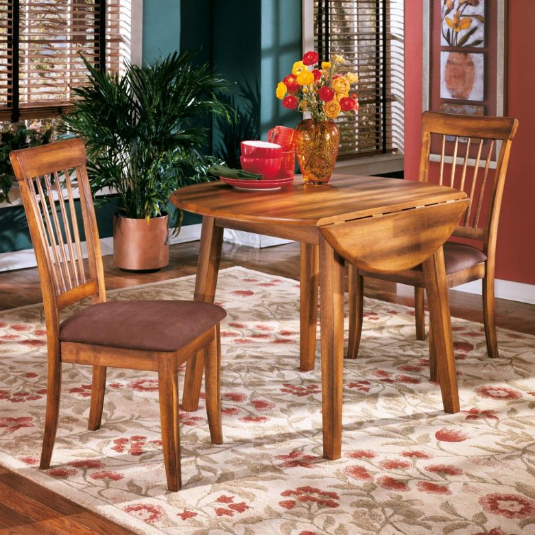 Cozy dining area with a wooden drop-leaf table, matching chairs, floral rug, and vase of flowers.