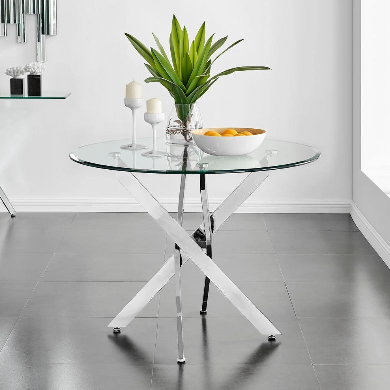 Round glass dining table with chrome crossed-leg base, decorated with a vase of green leaves, two white candles, and a bowl of oranges