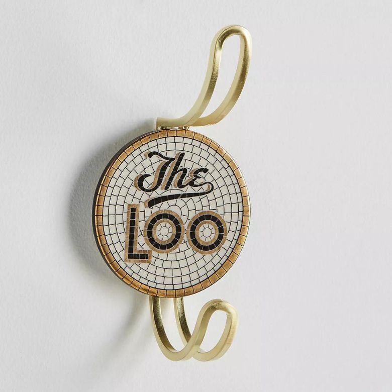Round wall hook with gold metal double hooks and a mosaic tile design featuring the words “The Loo” in black and gold lettering.