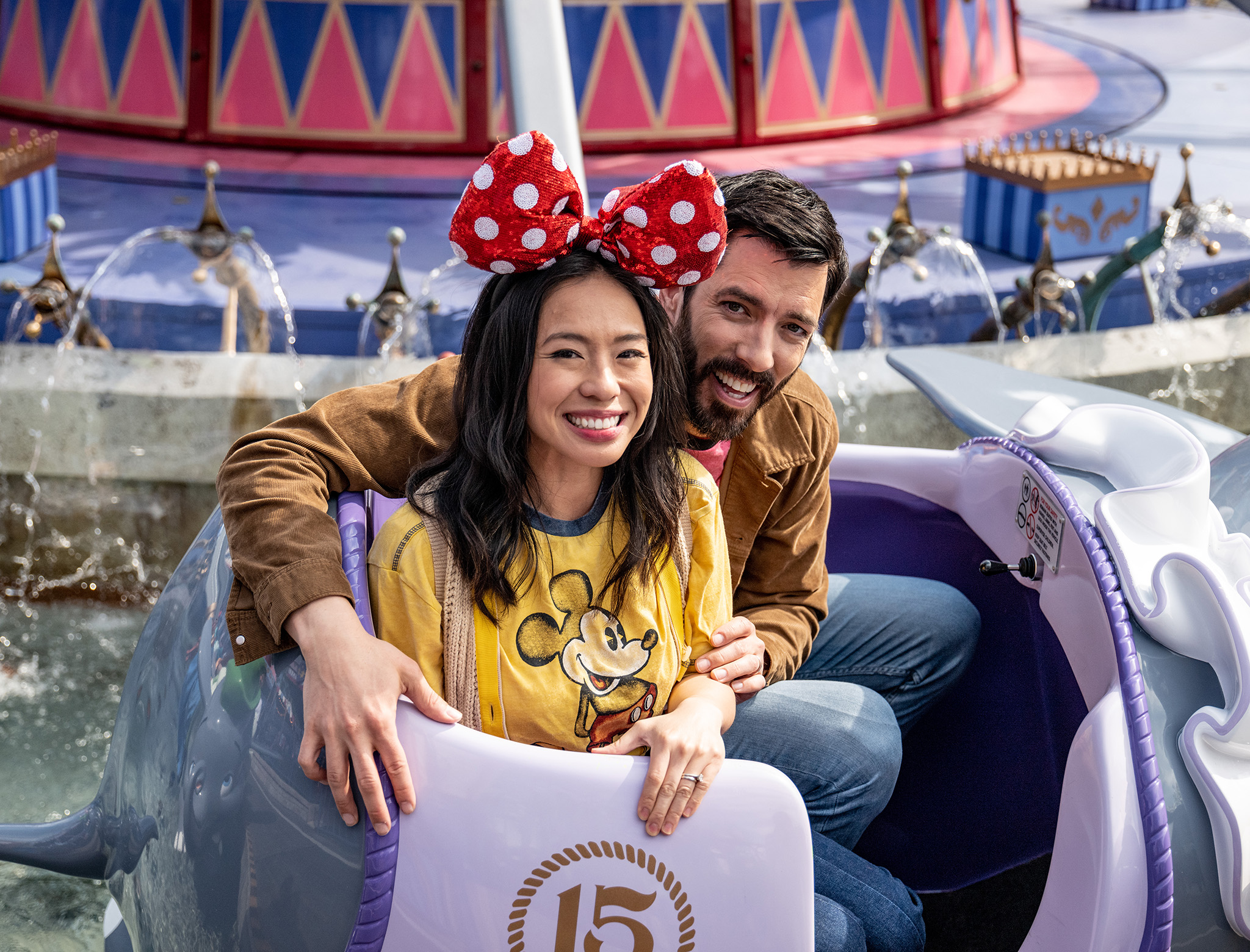 Smiling couple sitting together on a theme park ride with colorful decorations and fountains in the background.