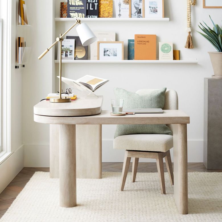 Light wood desk with a cream chair, books, and a brass lamp in a bright home office.
