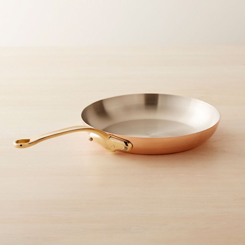 Copper frying pan with a polished interior and a gold-toned handle on a light wood surface.