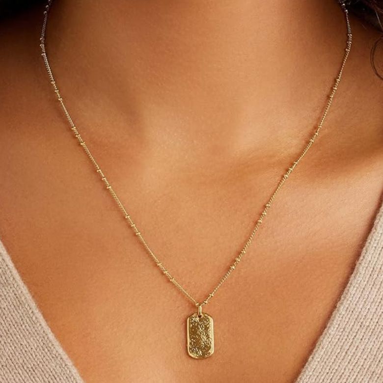 Gold pendant necklace on a delicate beaded chain worn against a beige top.