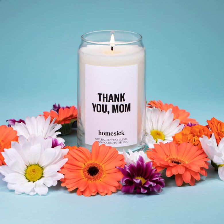 White “Thank You, Mom” candle surrounded by colorful daisies and gerbera flowers on a light blue background.