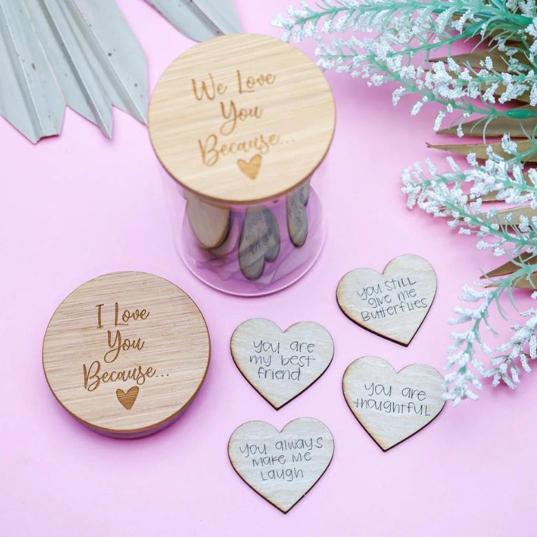 Wooden “I Love You Because” jars with engraved heart tokens laid out on a pink background.