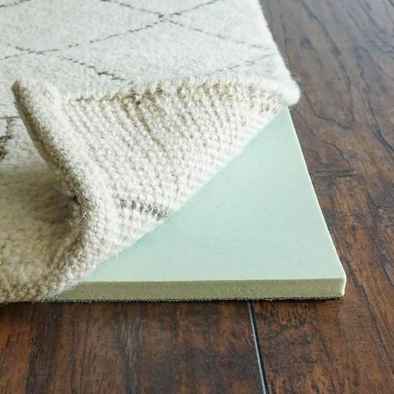 Corner of a textured rug lifted to show a thick foam rug pad beneath on a wood floor.