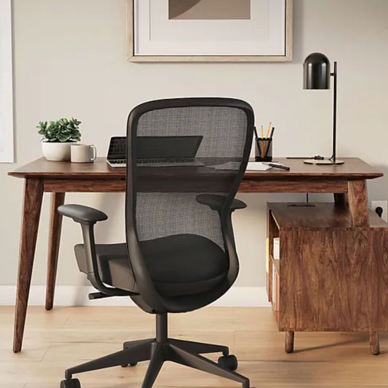Modern black mesh office chair at a wooden desk with a lamp, laptop, and decor.