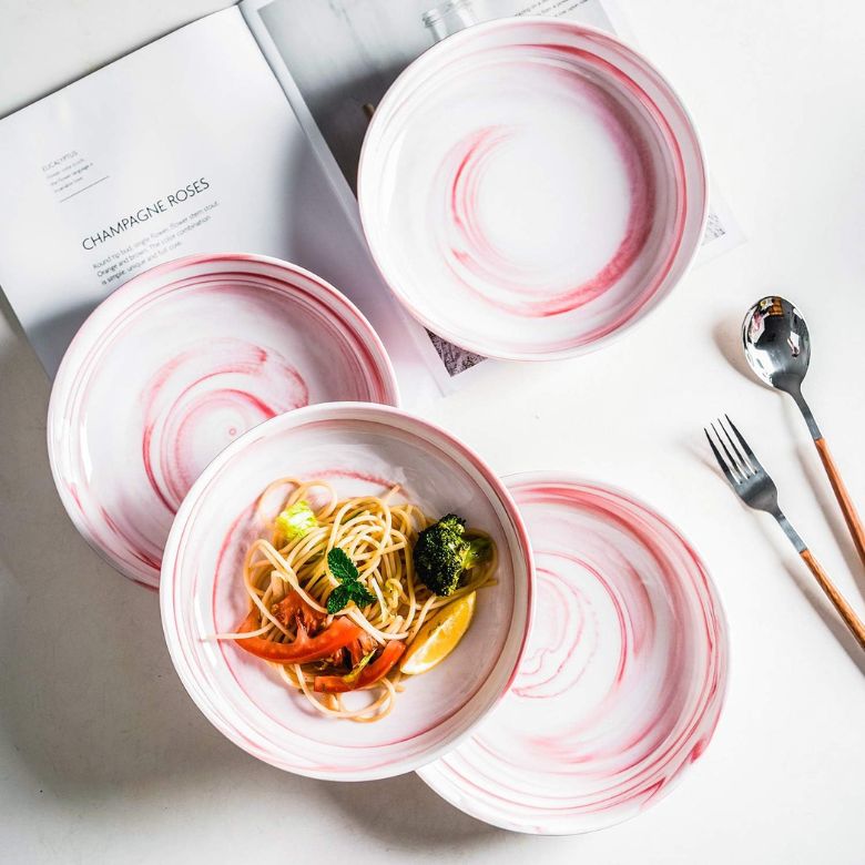Pink-swirled ceramic plates and bowls with pasta served in one bowl on a white table.
