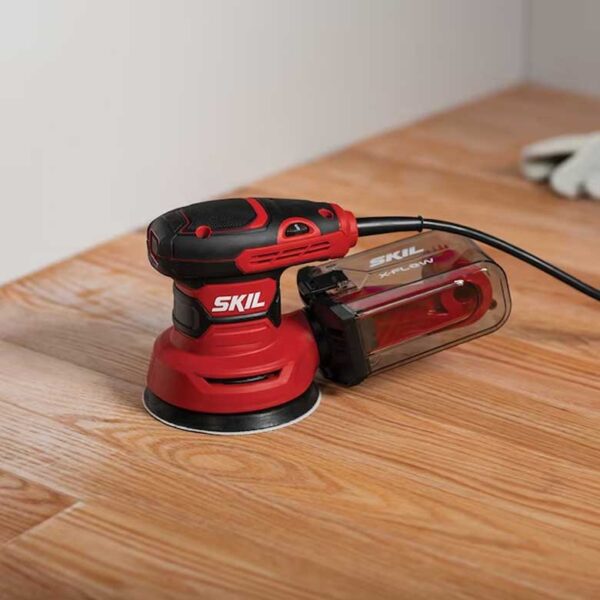 Red SKIL orbital sander placed on a wooden floor during a sanding project.