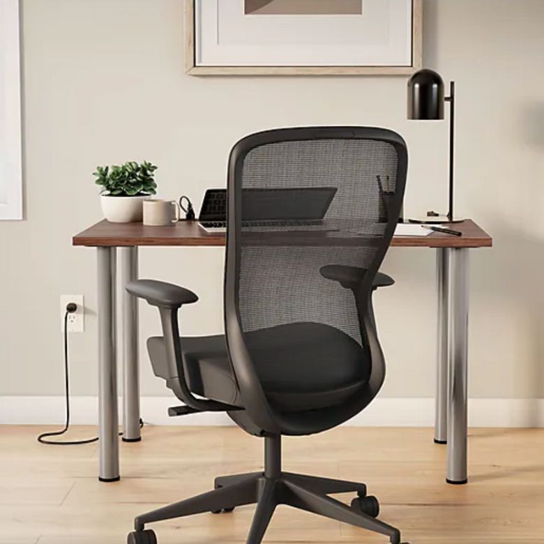 Home office setup with a black mesh desk chair facing a wooden desk with a lamp, laptop, and small plant.