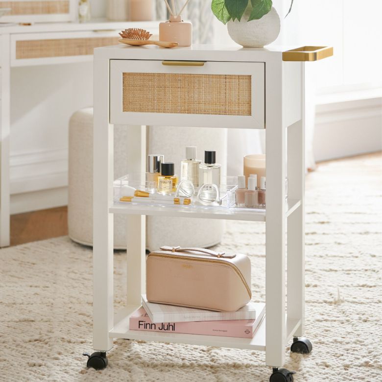 White rolling vanity cart with a woven-front drawer and shelves holding perfumes, makeup, and a cosmetic pouch.