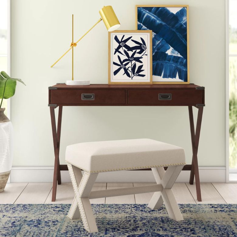 Wood console table with framed blue artwork and a gold lamp behind a cushioned bench on a patterned rug.