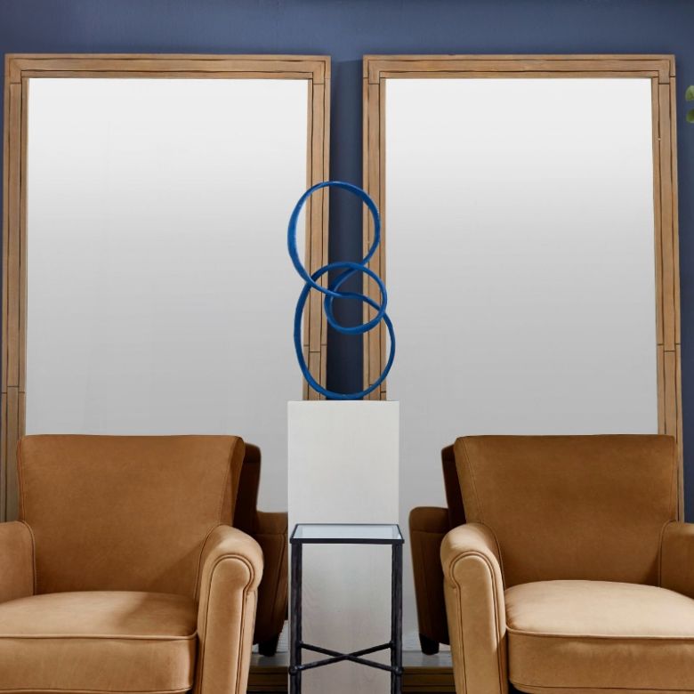 Two tan armchairs facing two large framed mirrors with a blue abstract sculpture on a pedestal between them.