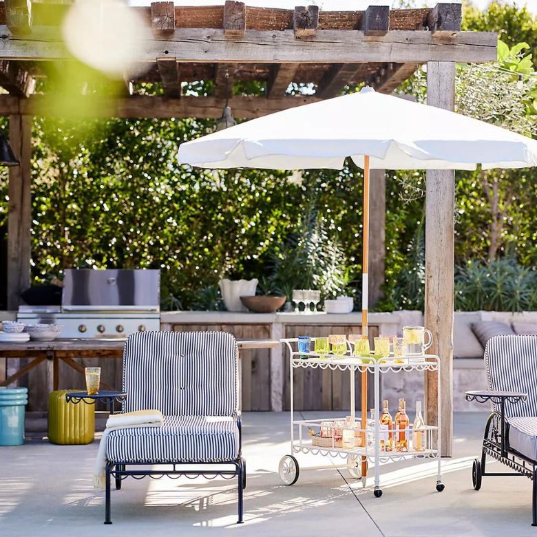Outdoor patio setup with striped lounge chairs, a white drink cart, and a large umbrella under a wooden pergola.