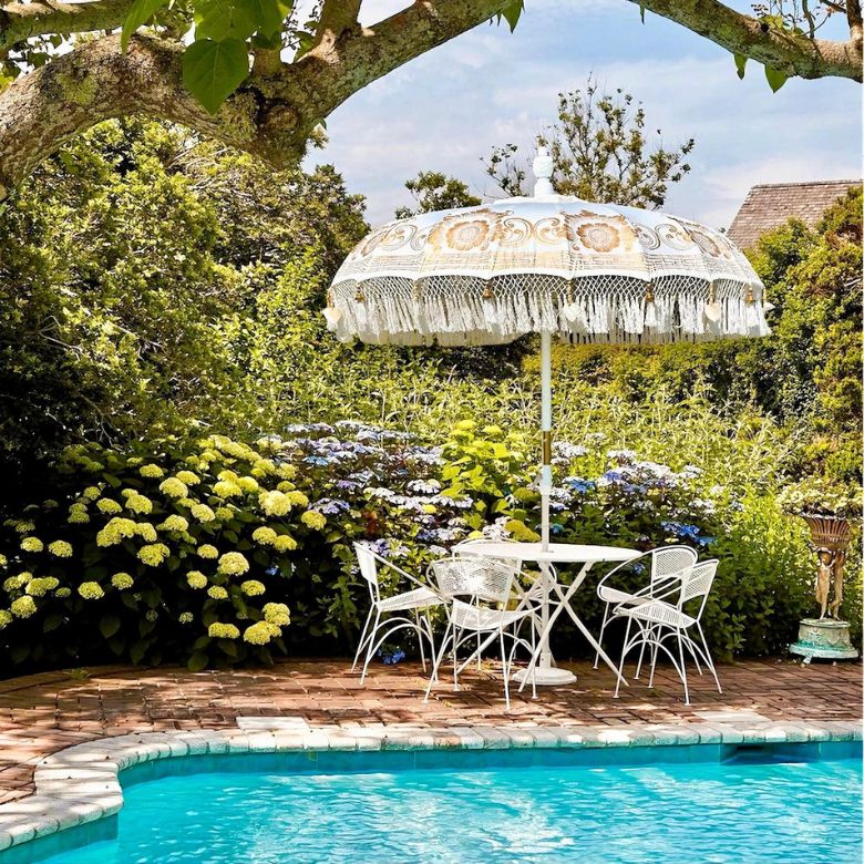Poolside seating area with a white fringe umbrella and metal chairs surrounded by lush blooming garden plants.