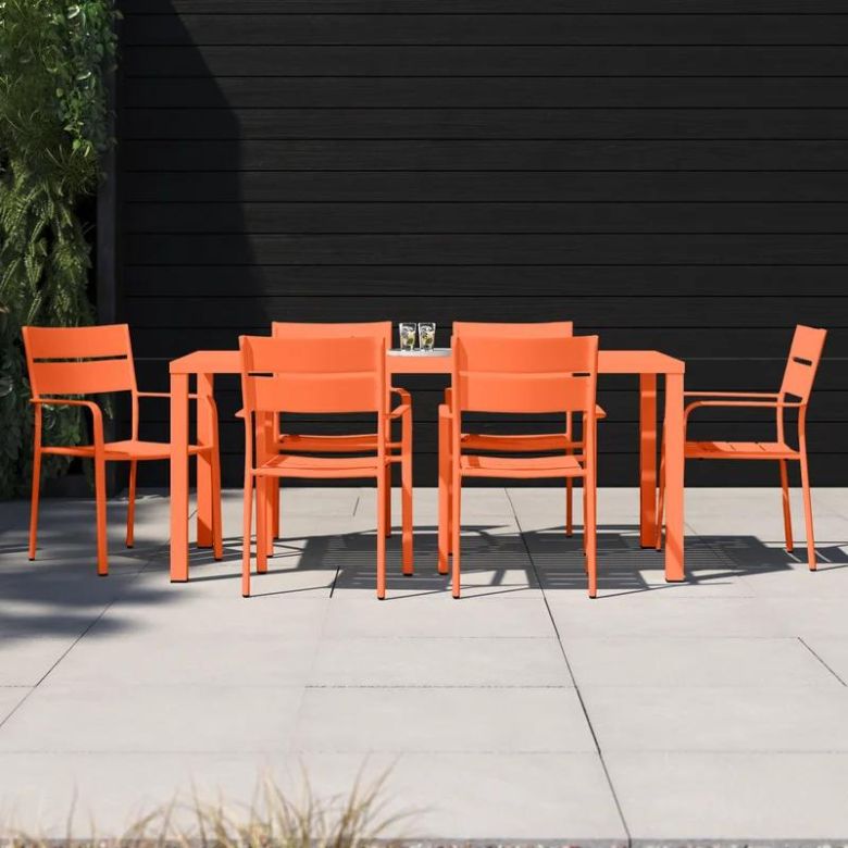 Bright orange outdoor dining table and chairs arranged on a light gray patio against a dark wood wall.