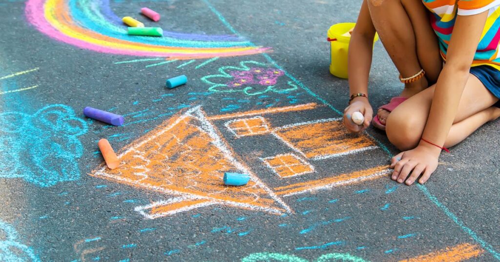 Child drawing colorful chalk art on pavement, including a house and a rainbow.