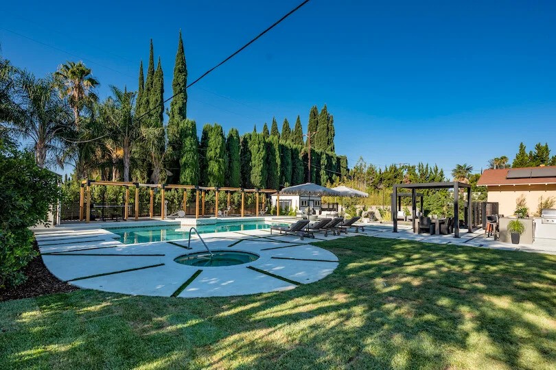Spacious backyard with pool, circular hot tub, lounge chairs, pergolas, and tall trees lining the fence
