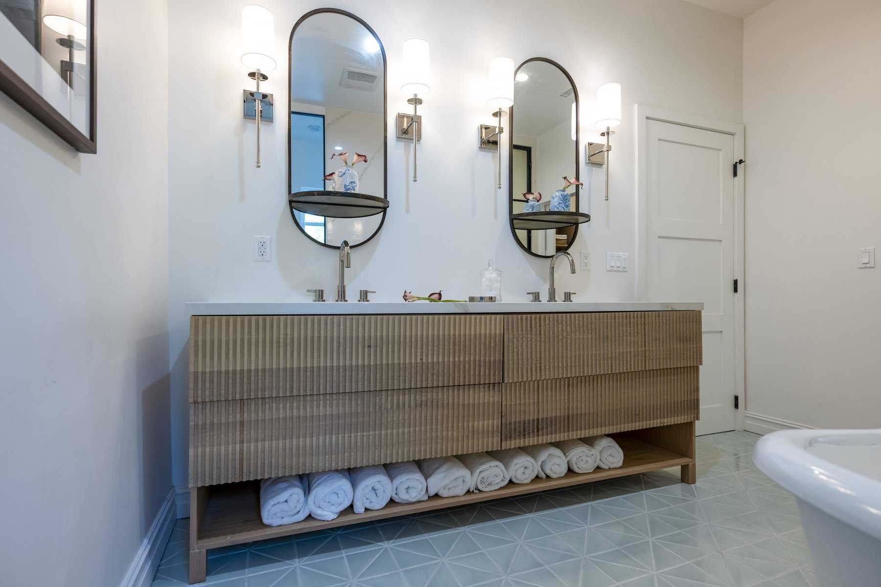 Modern bathroom with dual sinks, oval mirrors, wall sconces, and a wood vanity with rolled towels underneath
