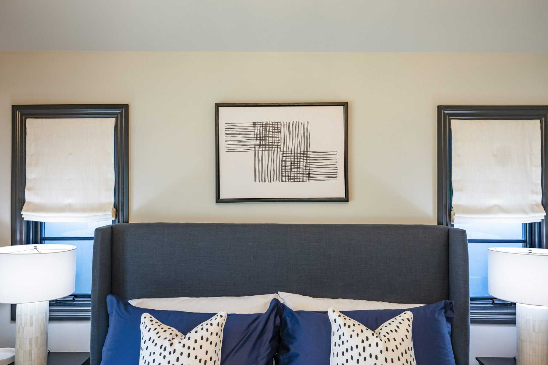 Bedroom with gray headboard, blue pillows, abstract art, and white Roman shades