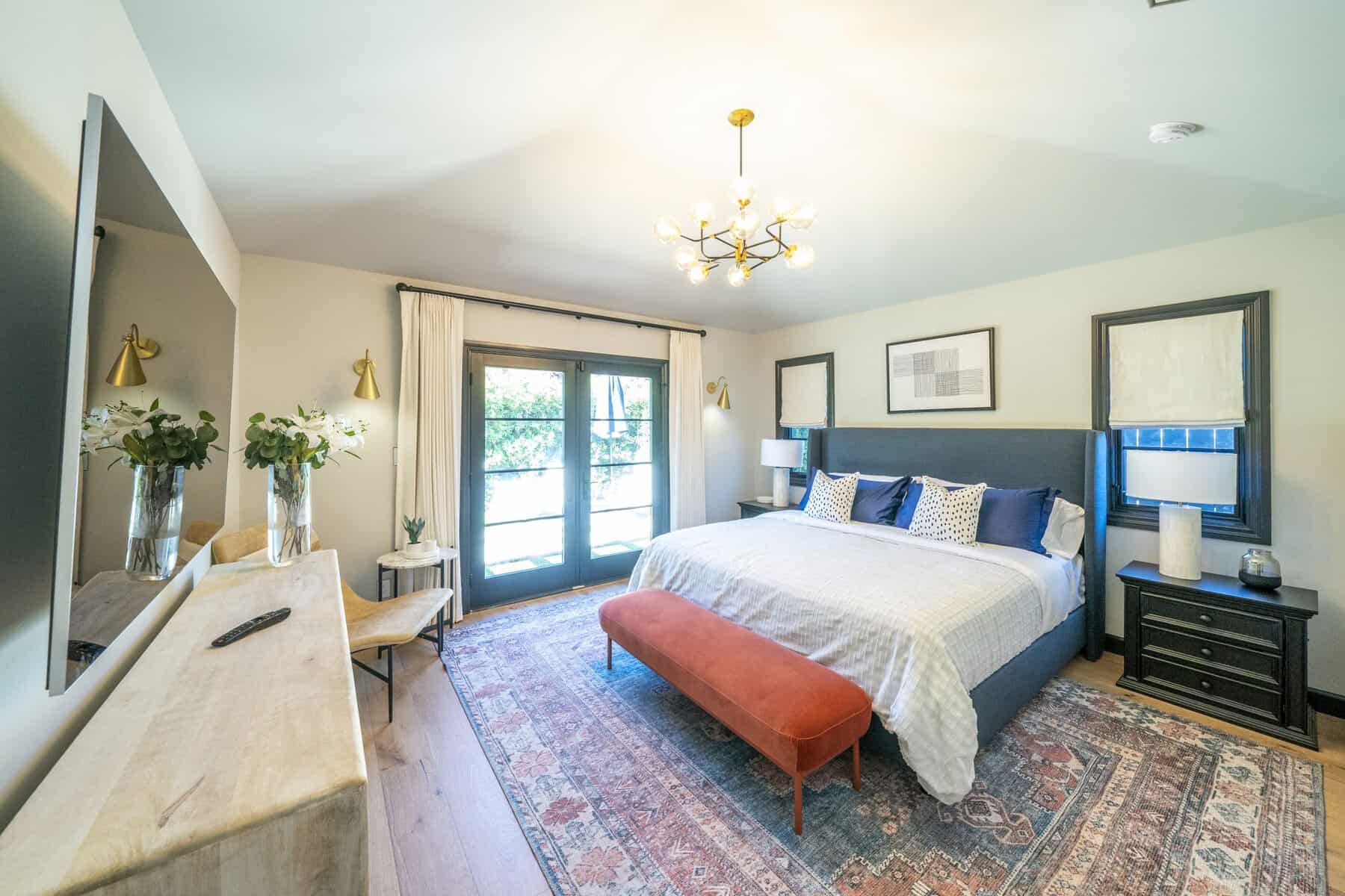 Bright bedroom with a patterned rug, navy upholstered bed, brass accents, and French doors framed by light curtains