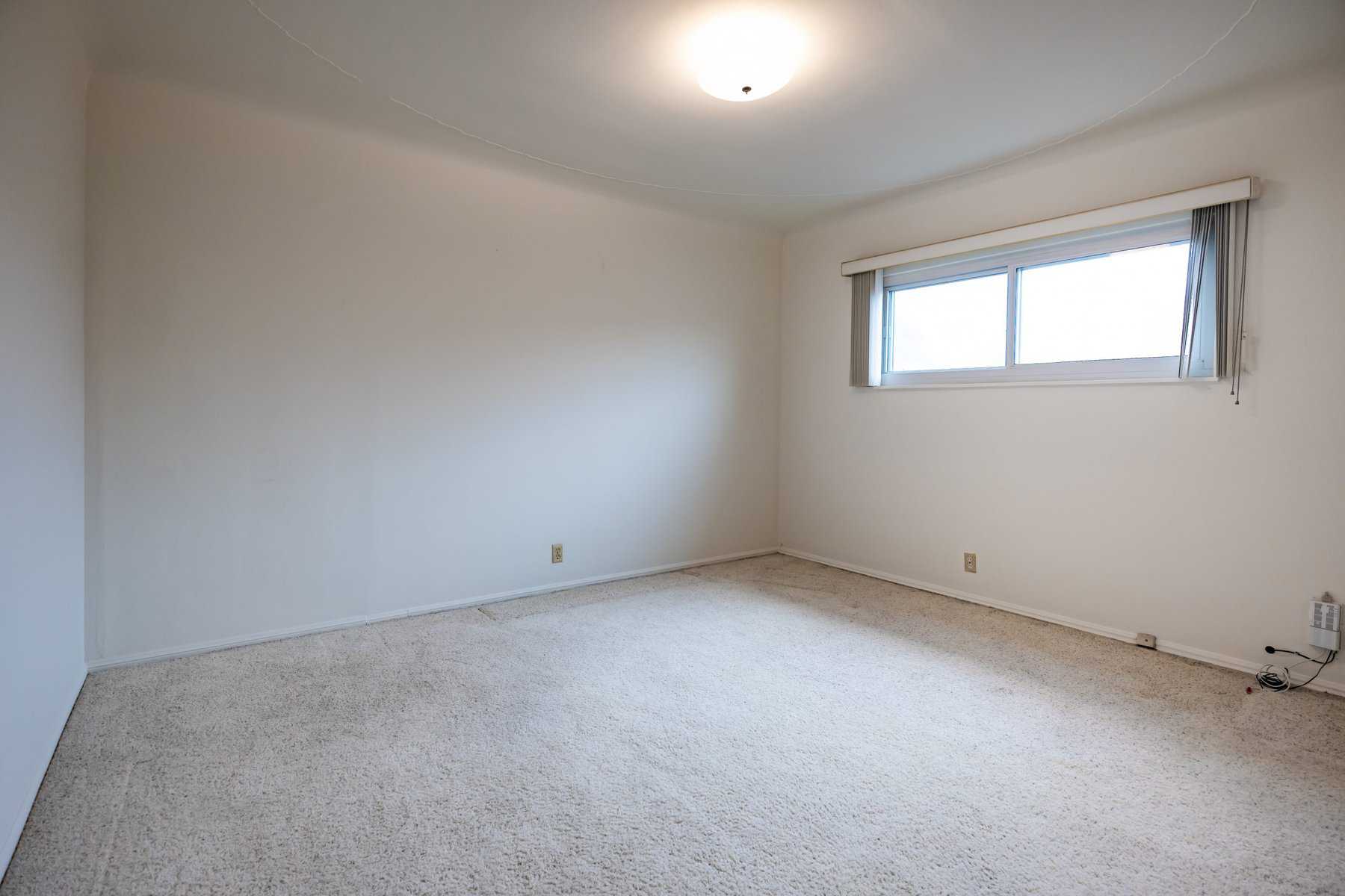 Empty bedroom with beige carpet, white walls, a ceiling light, and a large window with vertical blinds