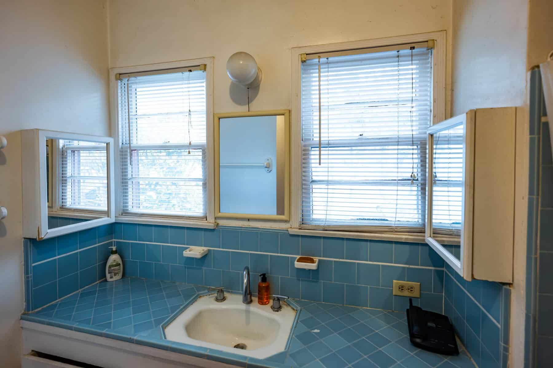 A bathroom with blue tiled counters, two windows with blinds, a mirror, and a light fixture above the sink