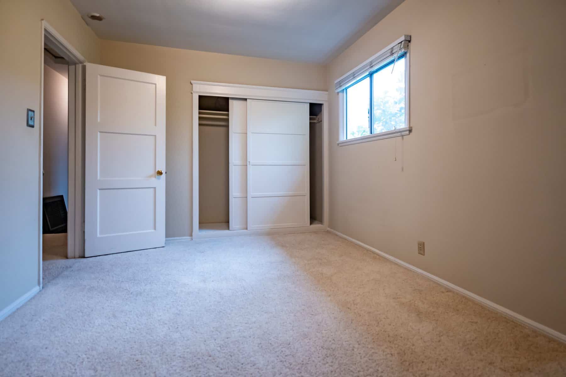 Empty bedroom with beige walls, white closet doors, carpet flooring, and a window letting in natural light