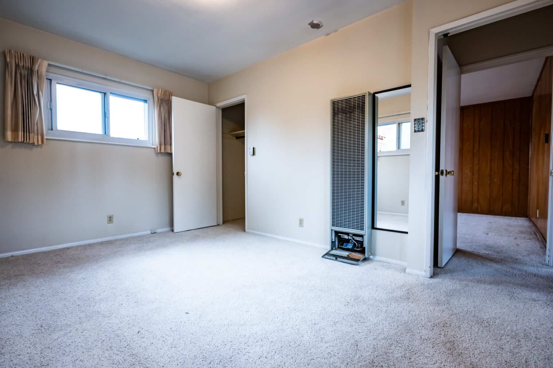 Empty carpeted room with beige walls, open doors, wall heater, and natural light from a window