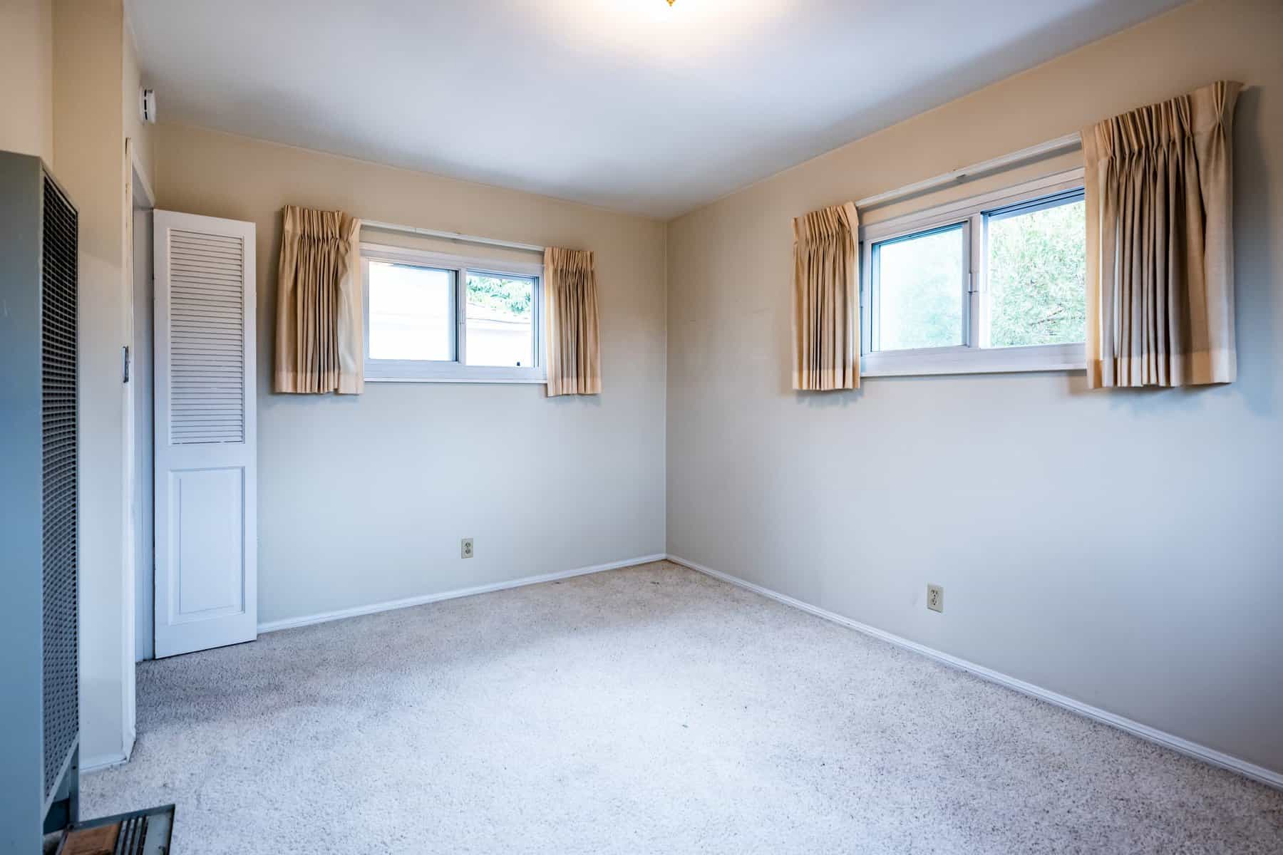 Empty room with beige walls carpeted floor and two windows with cream curtains