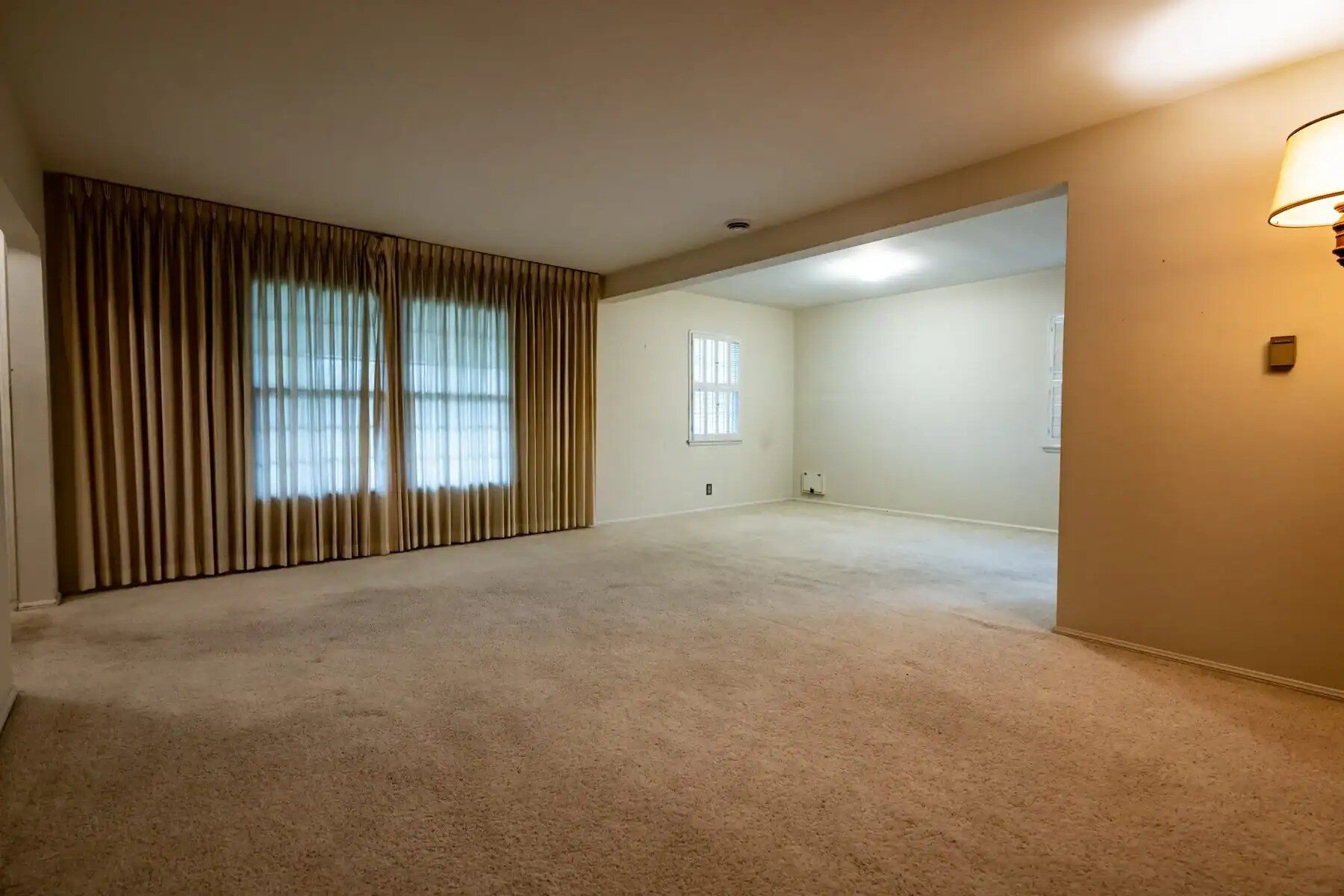 Empty living room with beige carpet, cream walls, and large windows with gold curtains