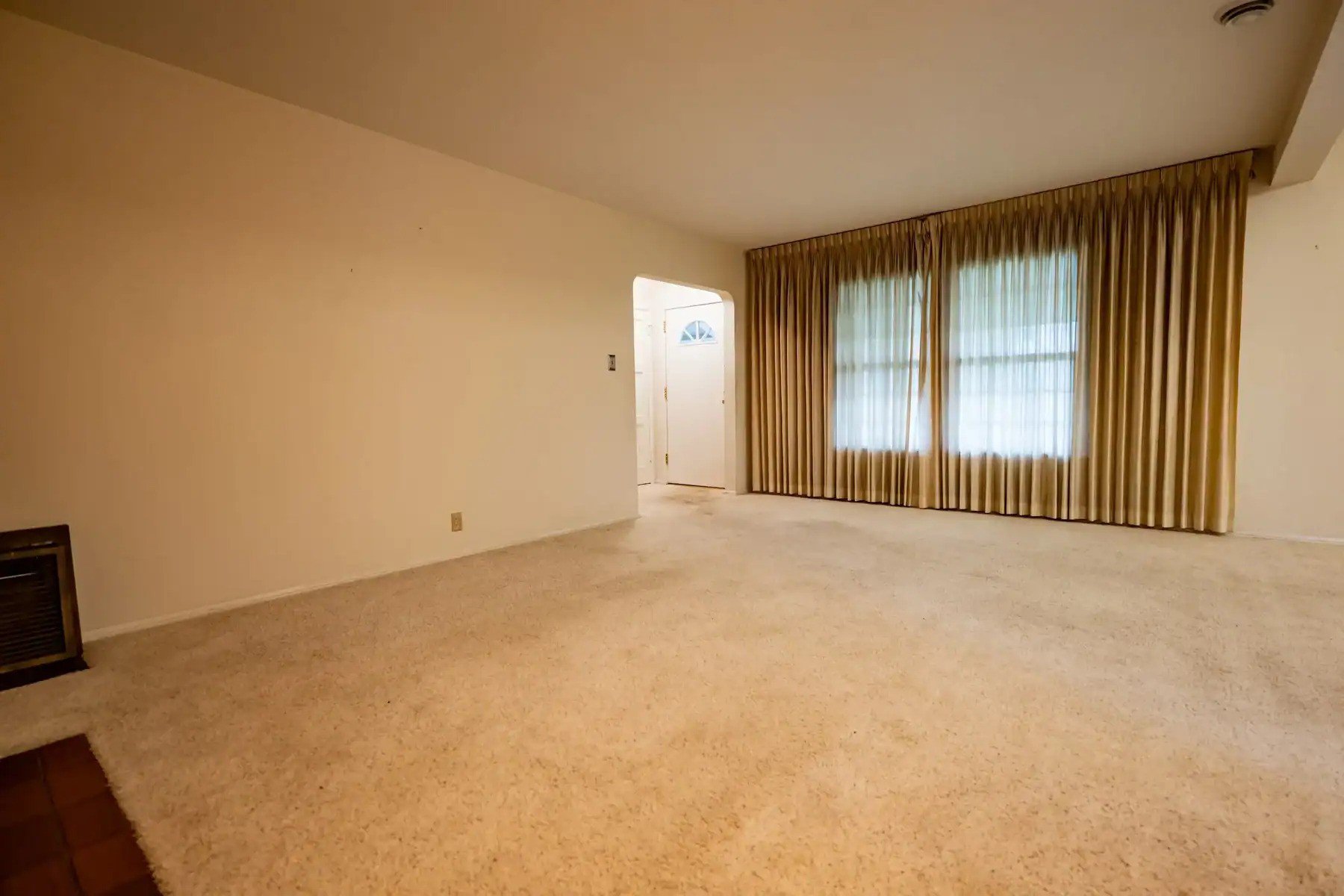 Empty living room with beige carpeting, gold curtains, and a doorway leading to the entrance