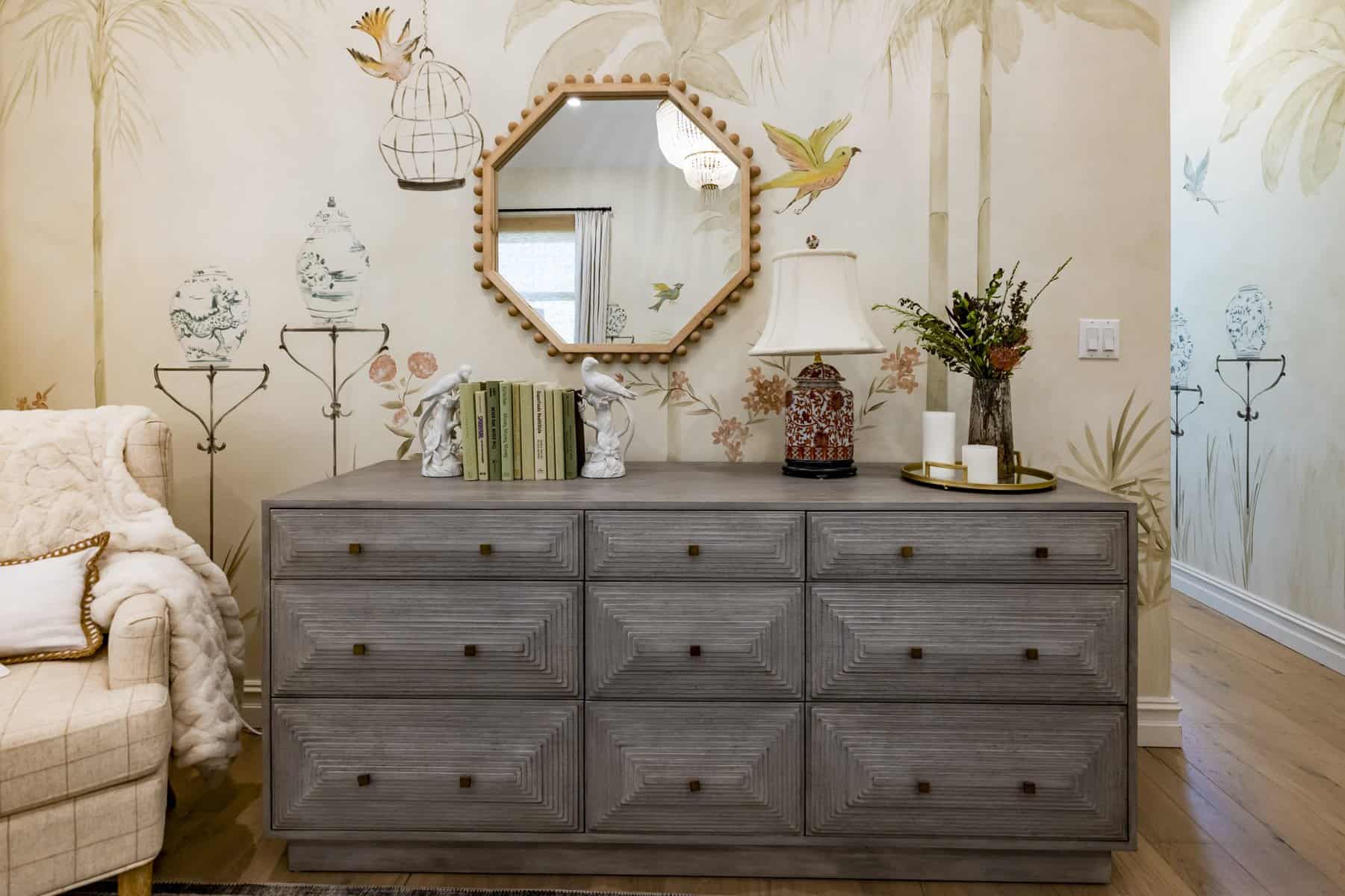 Gray wood dresser with octagonal mirror, floral wallpaper, books, and decorative table lamp