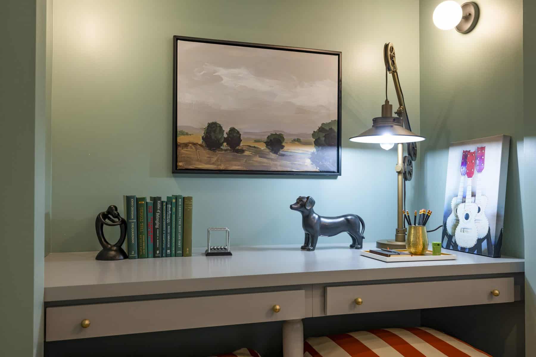 Cozy desk nook with a framed landscape painting, books, decorative sculptures, and a brass lamp against a muted green wall
