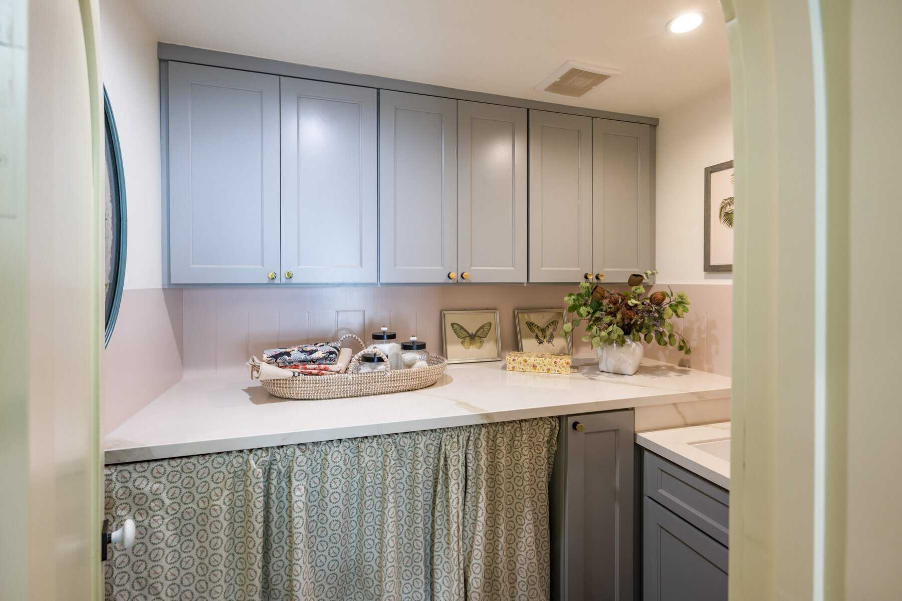 Light gray laundry room with cabinetry, countertop, decorative plants, and butterfly artwork