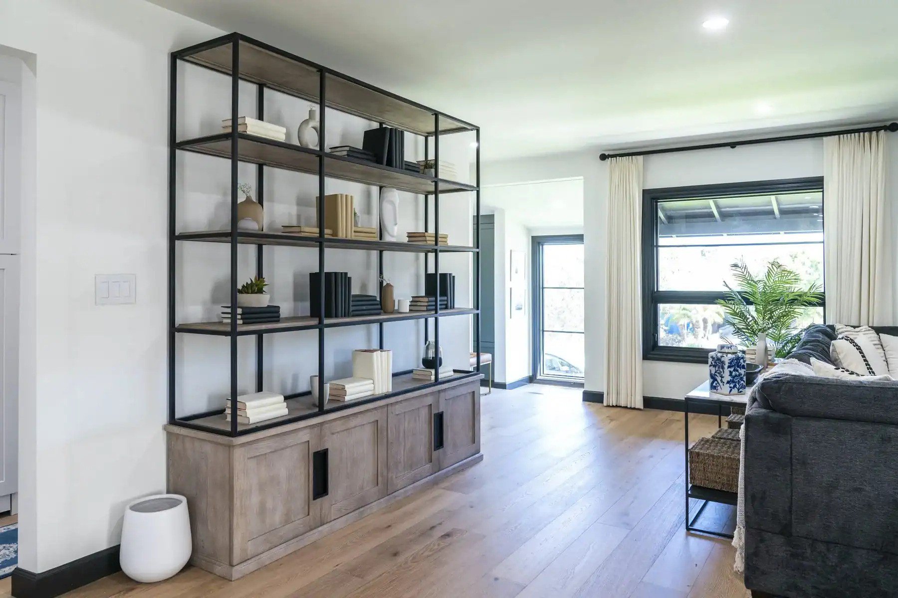 Living room with open shelving, light wood floors, and a large window with curtains