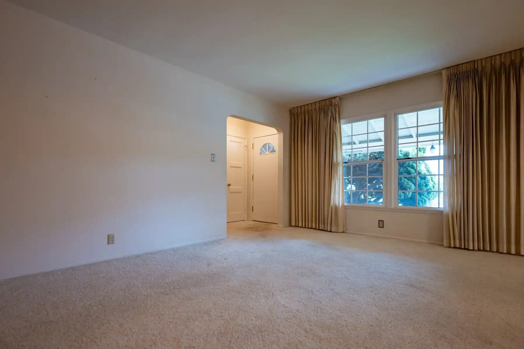 Empty living room with beige carpet, large window with drawn curtains, and an arched entryway