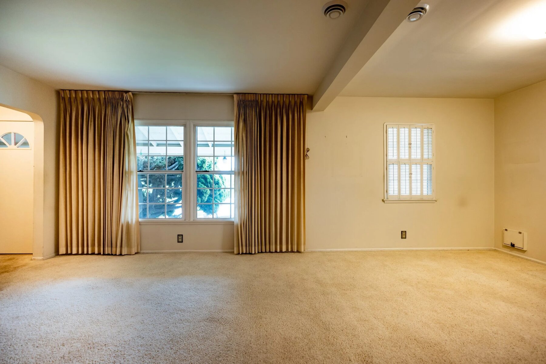 Empty living room with beige carpet, gold curtains, and two windows letting in natural light