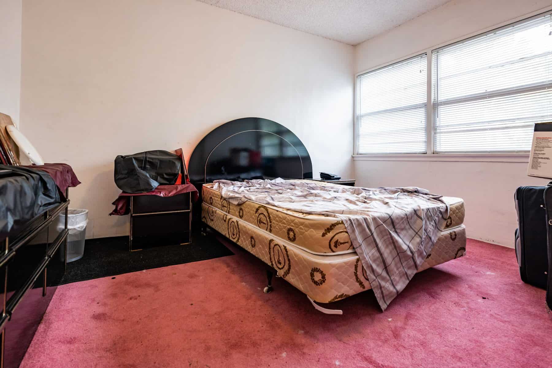 Cluttered bedroom with an unmade bed, mismatched furniture, and a pink carpet