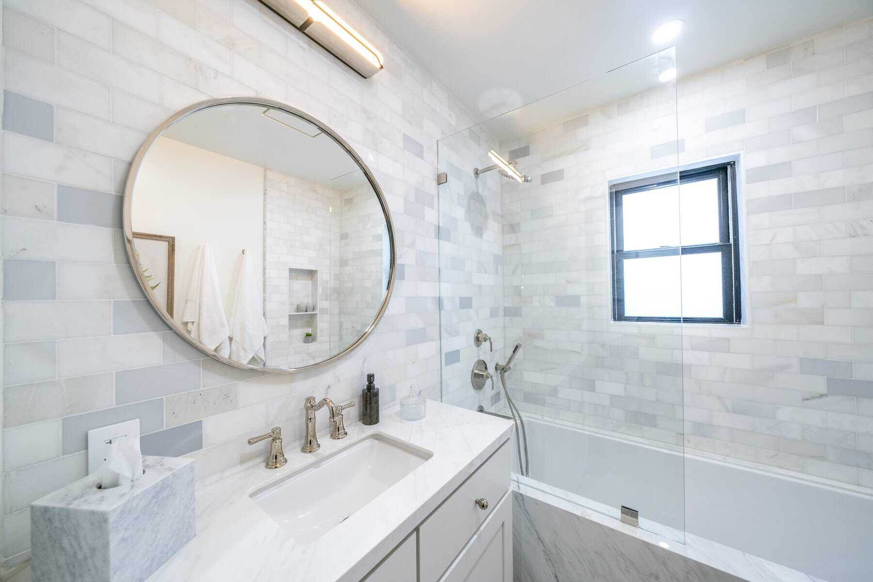Modern bathroom with white marble walls, a round mirror, glass shower panel, and sleek fixtures