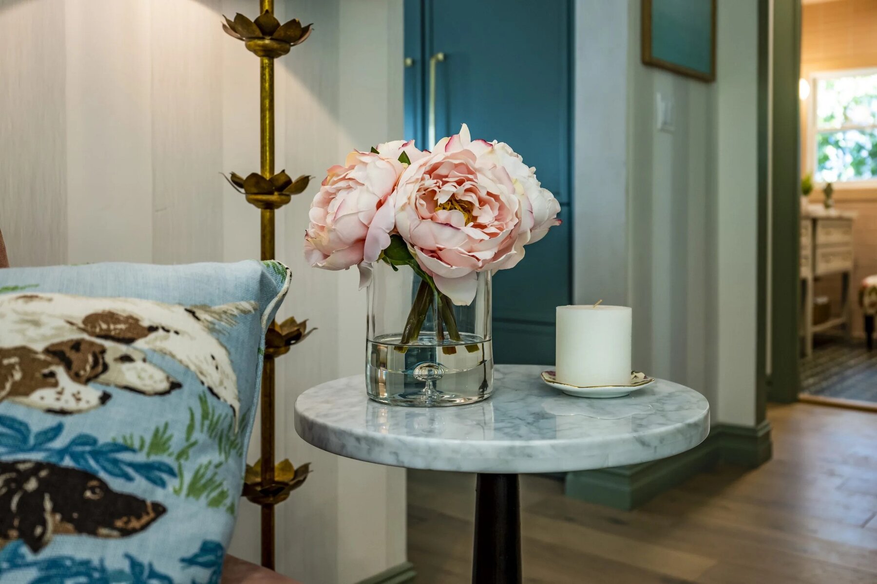 Marble side table with pink peonies in a glass vase, a white candle, and a decorative pillow nearby