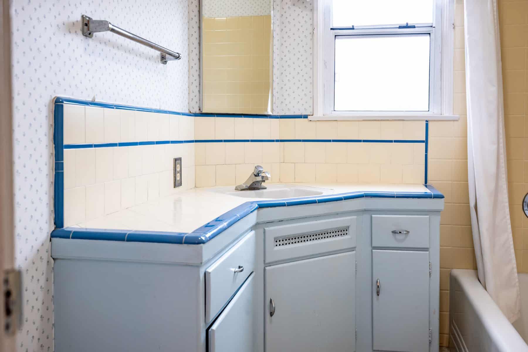 A vintage bathroom with light blue cabinets, cream tiles bordered in blue, a small sink, wallpaper, and a window above the countertop
