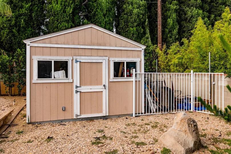 Tan backyard shed with white trim, gravel ground, and fenced side area storing tools