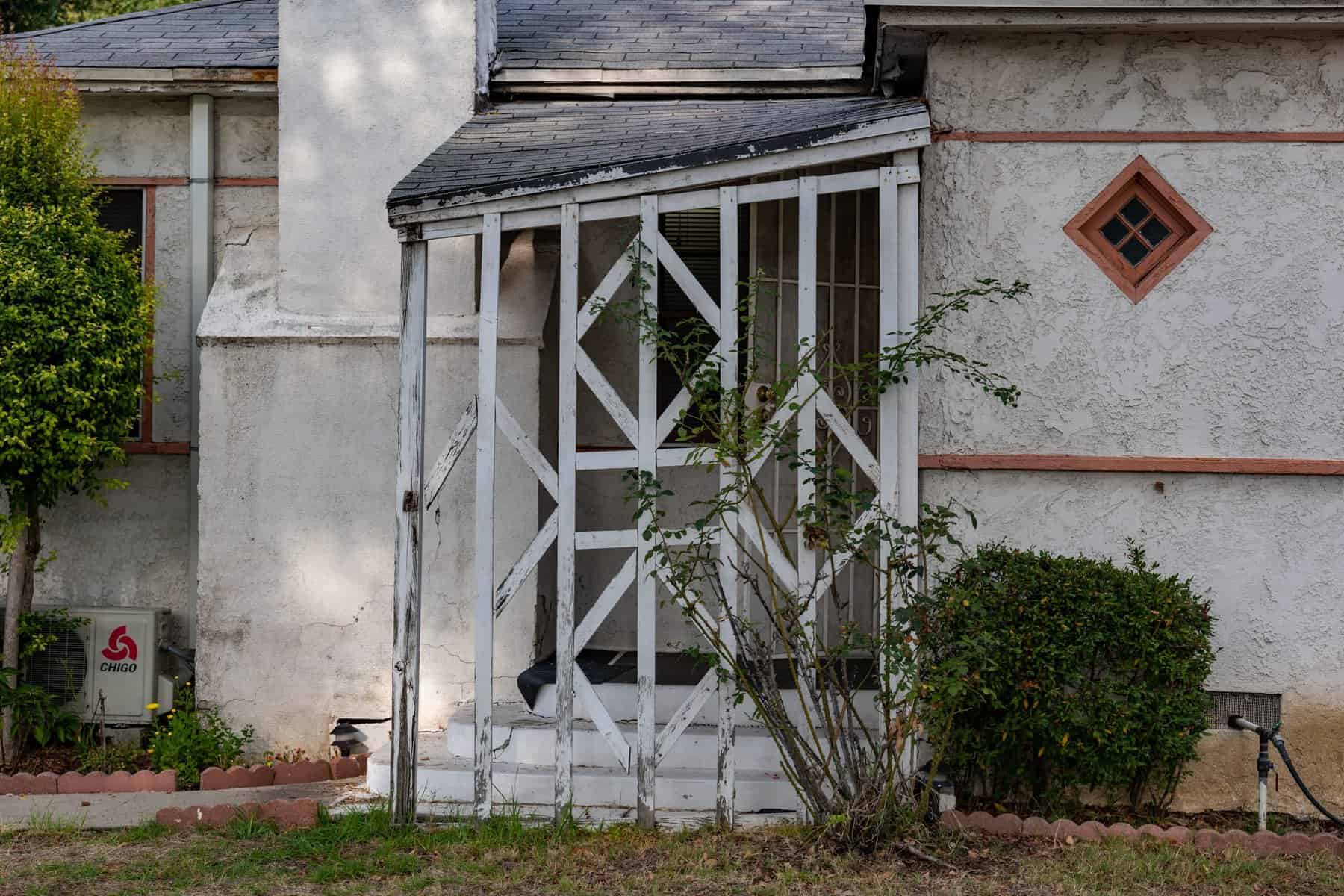 Weathered white stucco house with damaged porch, diamond-shaped window, sloping roof, chimney, shrubs, and trees