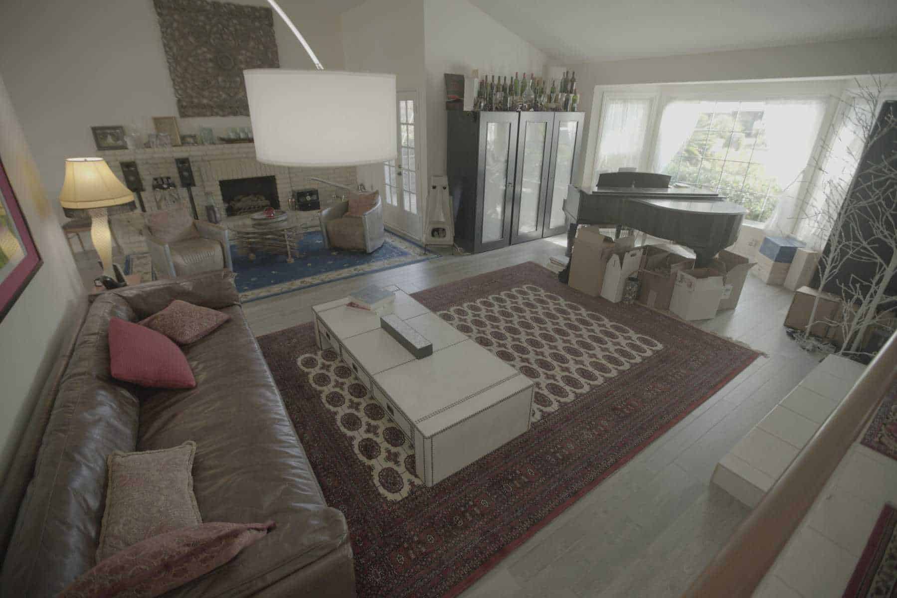 Cluttered living room with a leather sofa, patterned rugs, grand piano, and boxes near a large window