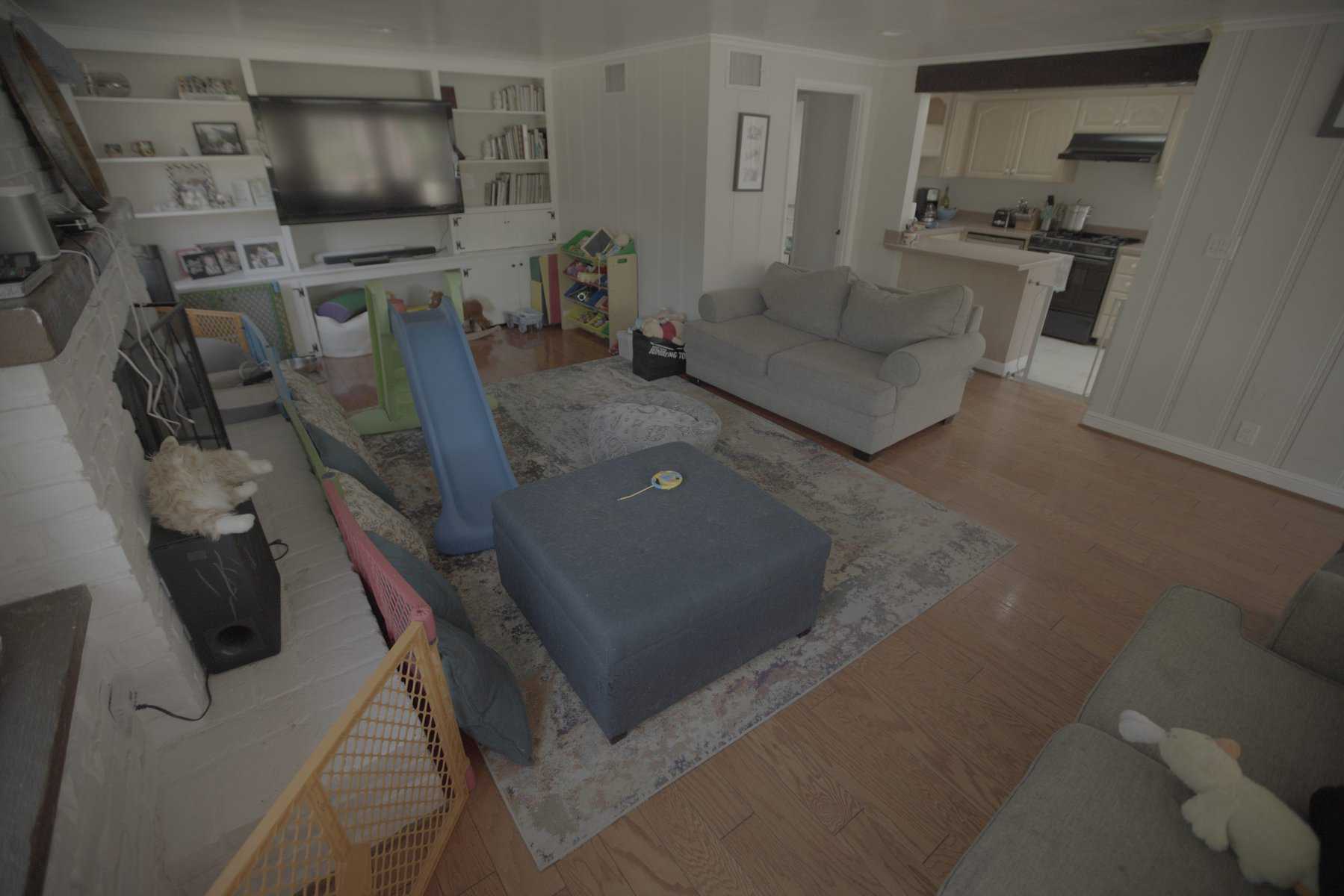 Cluttered living room with kids' slide, baby gates, toys, gray sofas, and open view to the kitchen