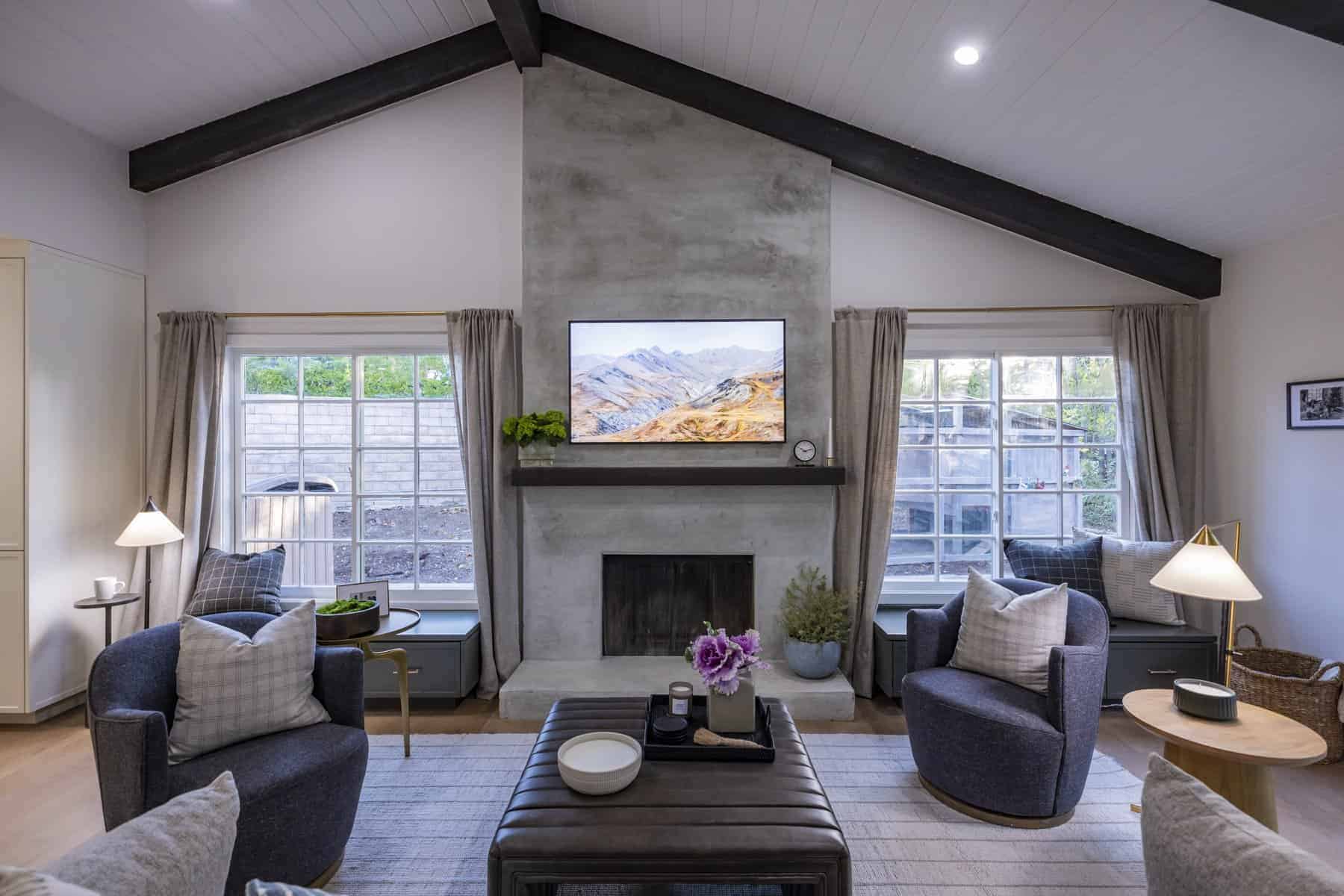 Cozy living room with vaulted ceiling, gray armchairs, a concrete fireplace wall with mounted TV, and symmetrical windows with light curtains