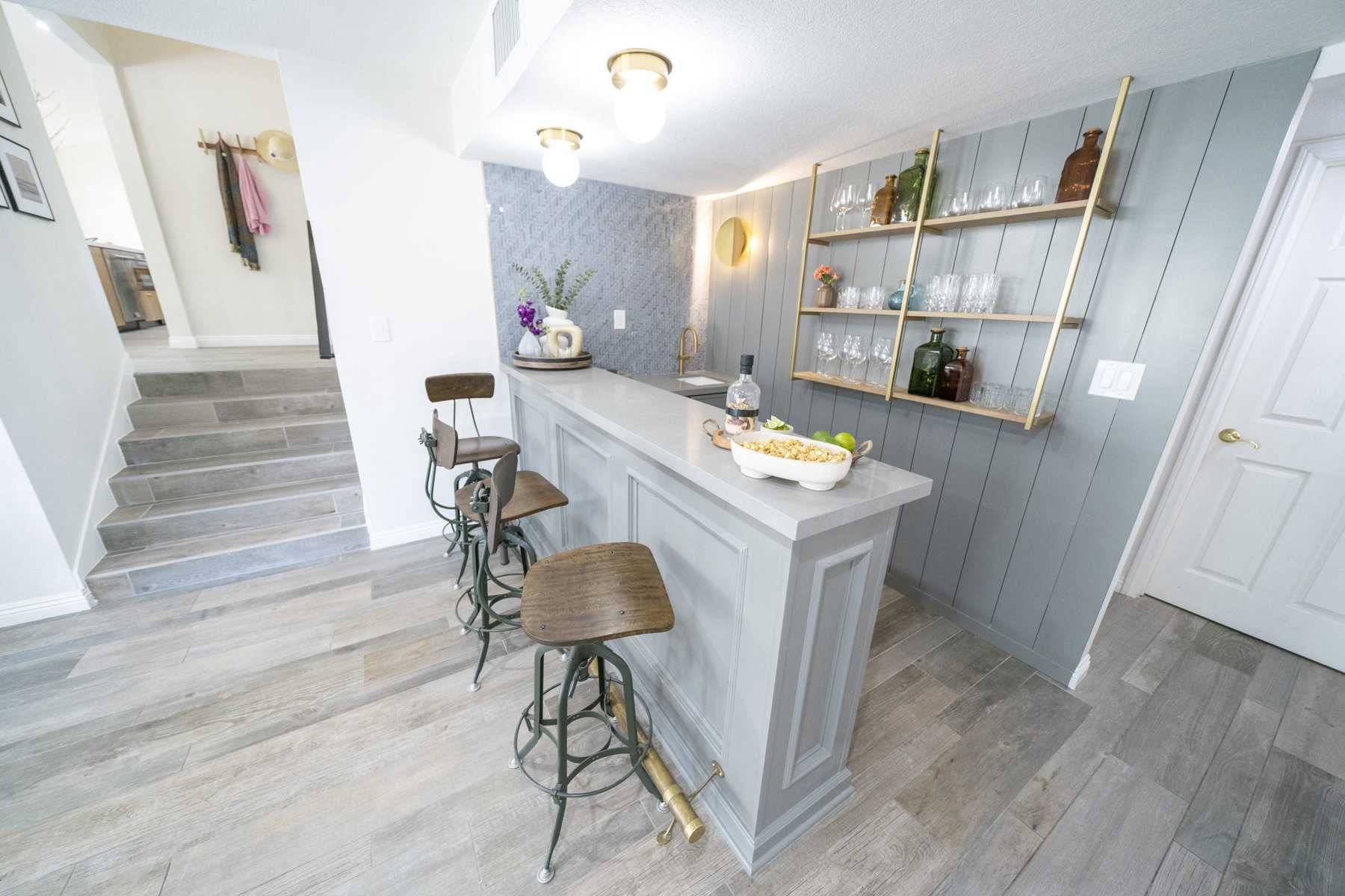 Modern home bar with gray paneling, open gold shelving, wooden bar stools, and decorative accents