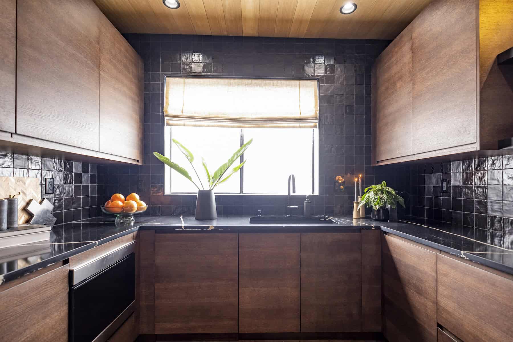Modern kitchen with dark cabinetry, black tiles, and a window with plants and a bowl of oranges on the countertop